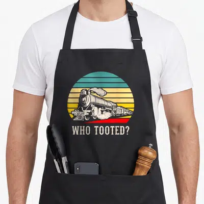Tooted locomotive railroad apron - Retro steam train in striped circle with text WHO TOOTED below — shown on a black cotton canvas apron against a white background