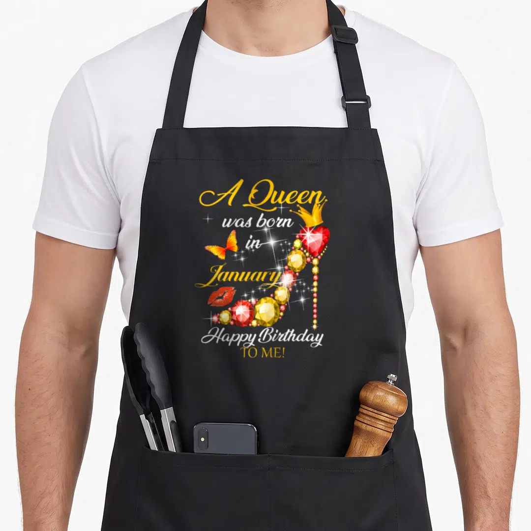 Funny queen january birthday apron - Jeweled high heel shoe with crown, red heart, butterfly, and lips surrounding gold — shown on a black cotton canvas apron against a white background