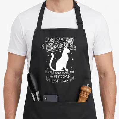 Witch cat apron - White cat silhouette with arched text reading Salem Sanctuary for Wayward Cats and Est 1692 — shown on a black cotton canvas apron against a white background