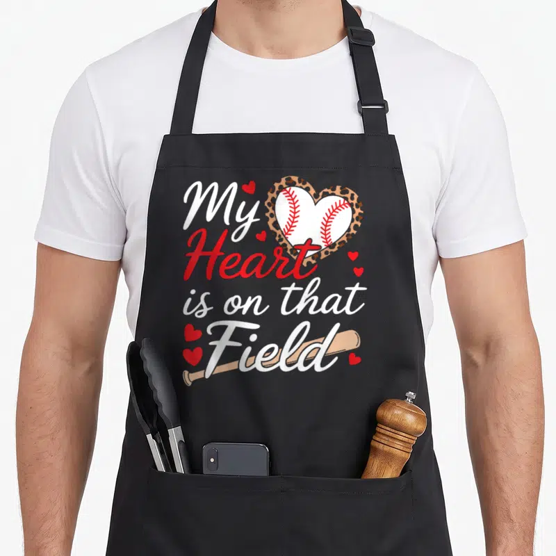 Field baseball grandma apron - My Heart is on that Field text with baseball in leopard heart and tan bat — shown on a black cotton canvas apron against a white background