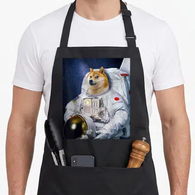 Shiba cosmonaut apron - Shiba Inu in white space suit holding gold helmet against starry blue background — shown on a black cotton canvas apron against a white background