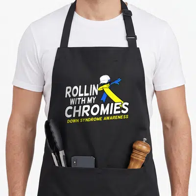 Syndrome little miss apron - Cartoon blue and yellow ribbon with cap next to ROLLIN WITH MY CHROMIES text — shown on a black cotton canvas apron against a white background