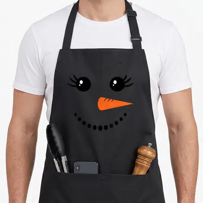 Snowman winter apron - Cartoon snowman face with eyelashes, orange nose, and black dot smile on grey background — shown on a black cotton canvas apron against a white background