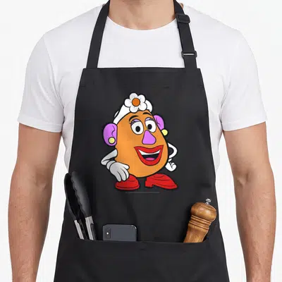 Funny potato mrs apron - Full body cartoon character with orange body, purple ears, white flower hat, and red shoes — shown on a black cotton canvas apron against a white background
