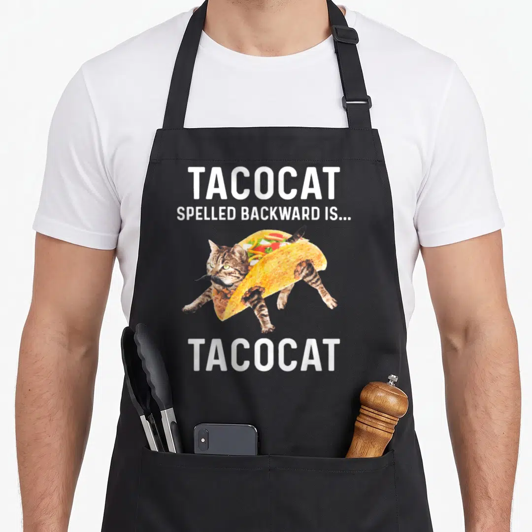 Tacocat taco apron - Cartoon cat inside a taco shell with white text reading TACOCAT spelled backward is Tacocat — shown on a black cotton canvas apron against a white background