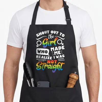 Shout gay pride apron - Stacked rainbow text reading Shout Out To The Girl Who Made Me Realize I Was Not Straight — shown on a black cotton canvas apron against a white background