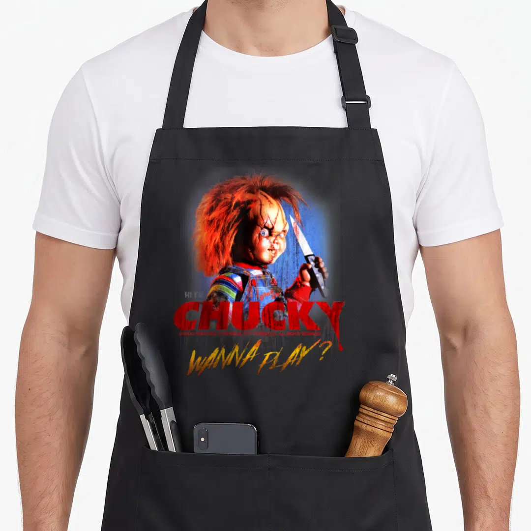 Funny play halloween apron - Chucky doll holding knife with red CHUCKY and yellow WANNA PLAY text — shown on a black cotton canvas apron against a white background