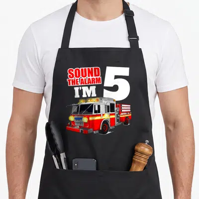 Five truck apron - Cartoon fire truck below Sound The Alarm I'm 5 text — shown on a black cotton canvas apron against a white background