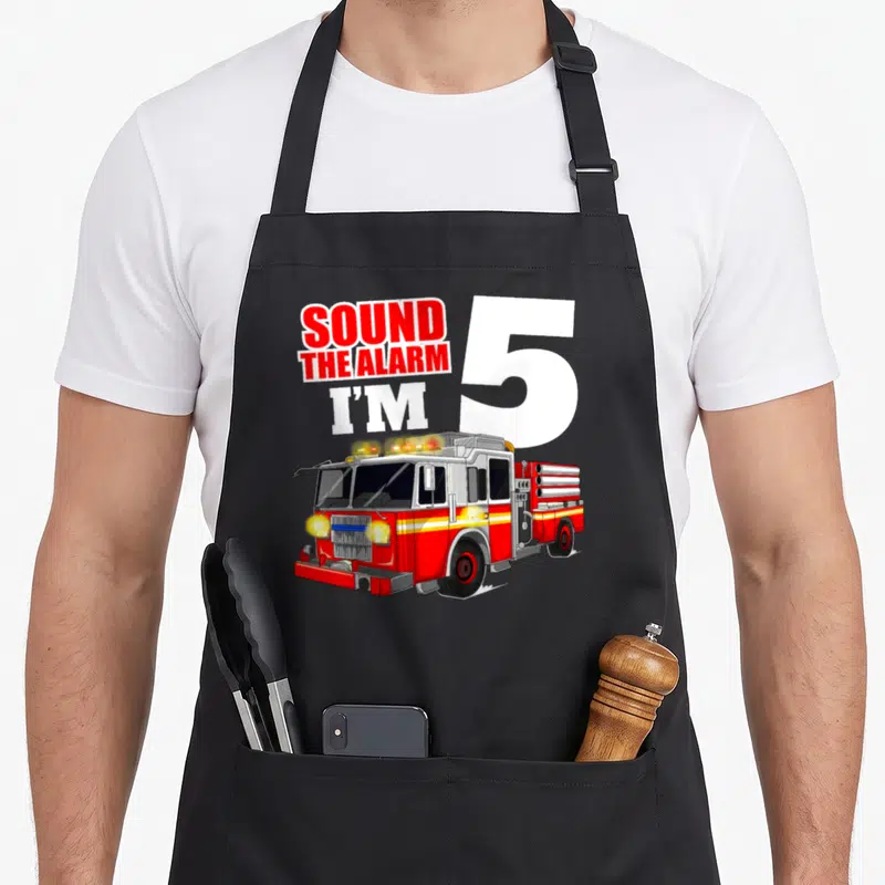 Five truck apron - Cartoon fire truck below Sound The Alarm I'm 5 text — shown on a black cotton canvas apron against a white background
