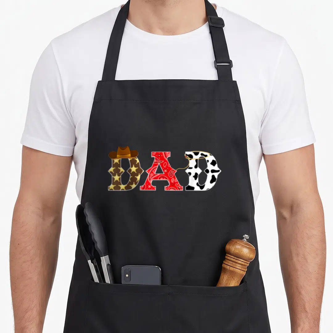 Theme western aprons - DAD text with cowboy hat, lasso, stars, paisley, and cow print pattern — shown on a black cotton canvas apron against a white background