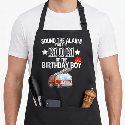 Sound truck apron - Fire truck and sound the alarm mom of birthday boy text with helmet and stars — shown on a black cotton canvas apron against a white background