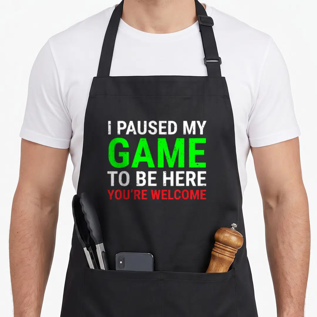 Paused gaming apron - Stacked text I paused my game to be here you're welcome in white green and red — shown on a black cotton canvas apron against a white background