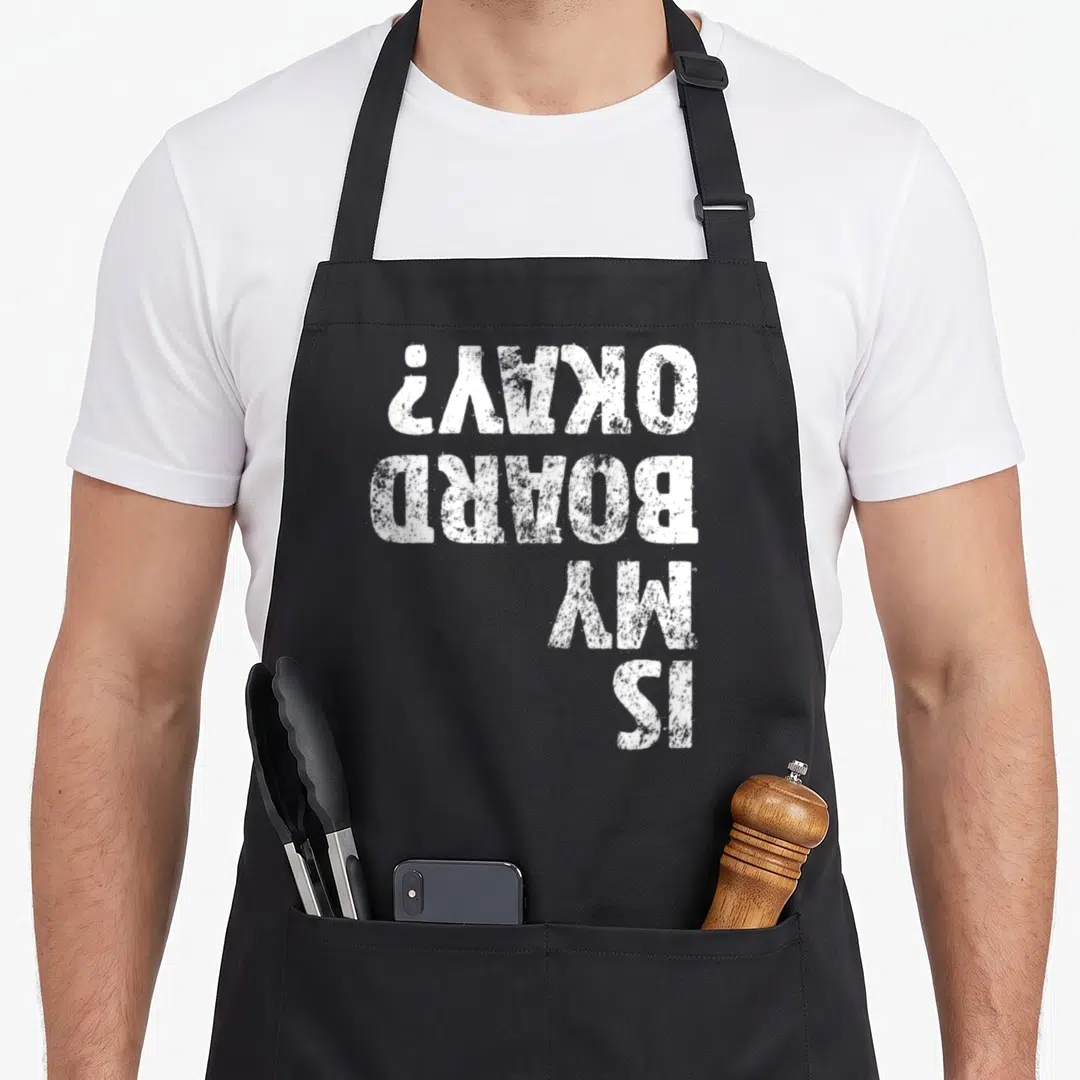 Board skateboarding apron - White distressed text stacked vertically reading IS MY BOARD OKAY on dark background — shown on a black cotton canvas apron against a white background