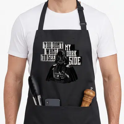 Funny dark novelty apron - Darth Vader holding remote surrounded by two text blocks reading YOU DON'T WANT TO SEE MY — shown on a black cotton canvas apron against a white background