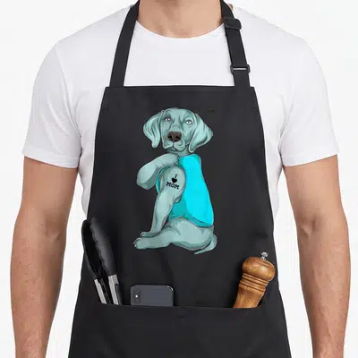 Cute weimaraner dog apron - Grey cartoon dog in cyan shirt showing I heart MOM tattoo on arm — shown on a black cotton canvas apron against a white background