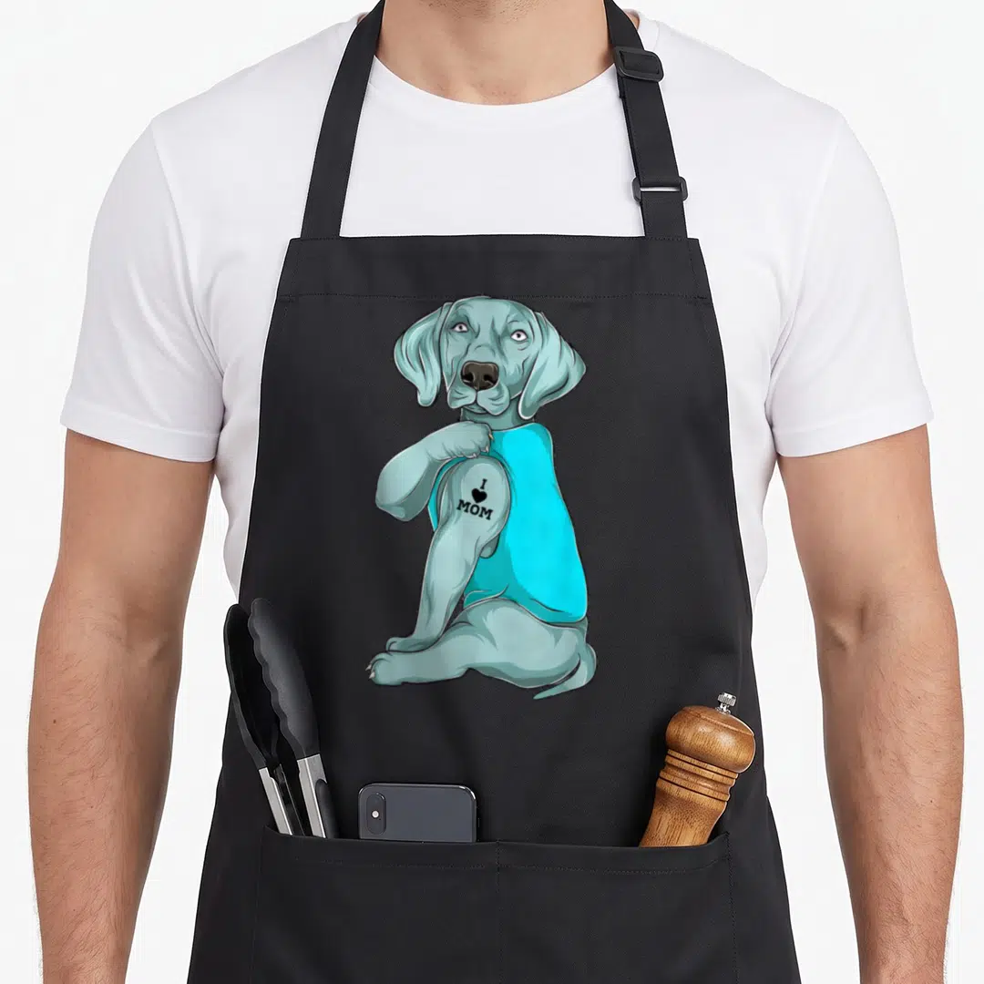 Cute weimaraner dog apron - Grey cartoon dog in cyan shirt showing I heart MOM tattoo on arm — shown on a black cotton canvas apron against a white background