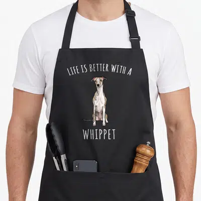 Better whippet dog apron - Sitting cartoon whippet in white tan and brown with LIFE IS BETTER WITH A WHIPPET text — shown on a black cotton canvas apron against a white background