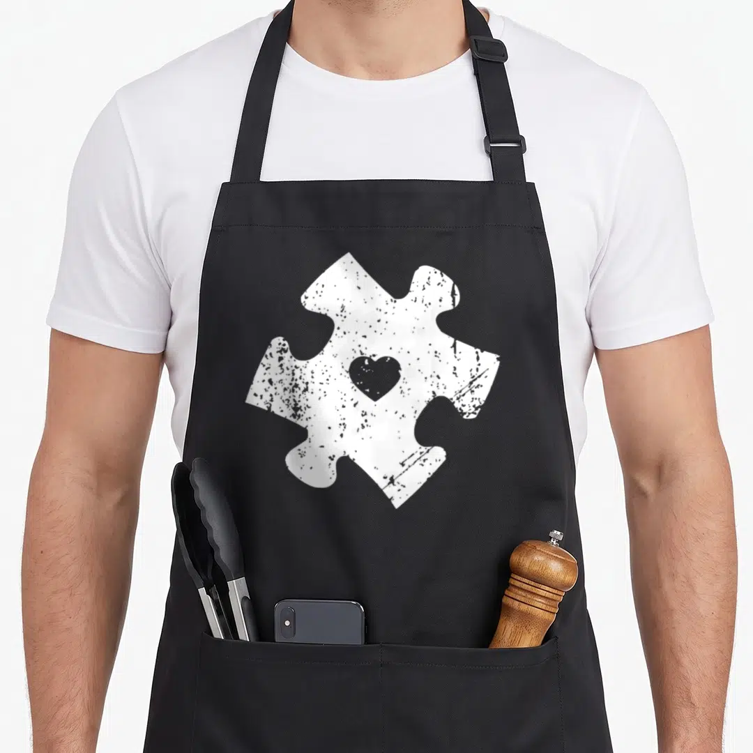 Autism teacher dad apron - Distressed white puzzle piece with grey heart cutout centered on front — shown on a black cotton canvas apron against a white background