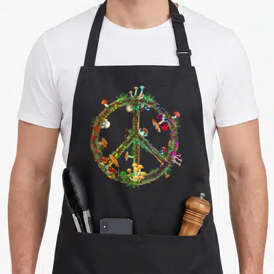 Magic mushroom apron - Brown peace sign made of mushrooms and grass with red, blue, and green paint splatters — shown on a black cotton canvas apron against a white background