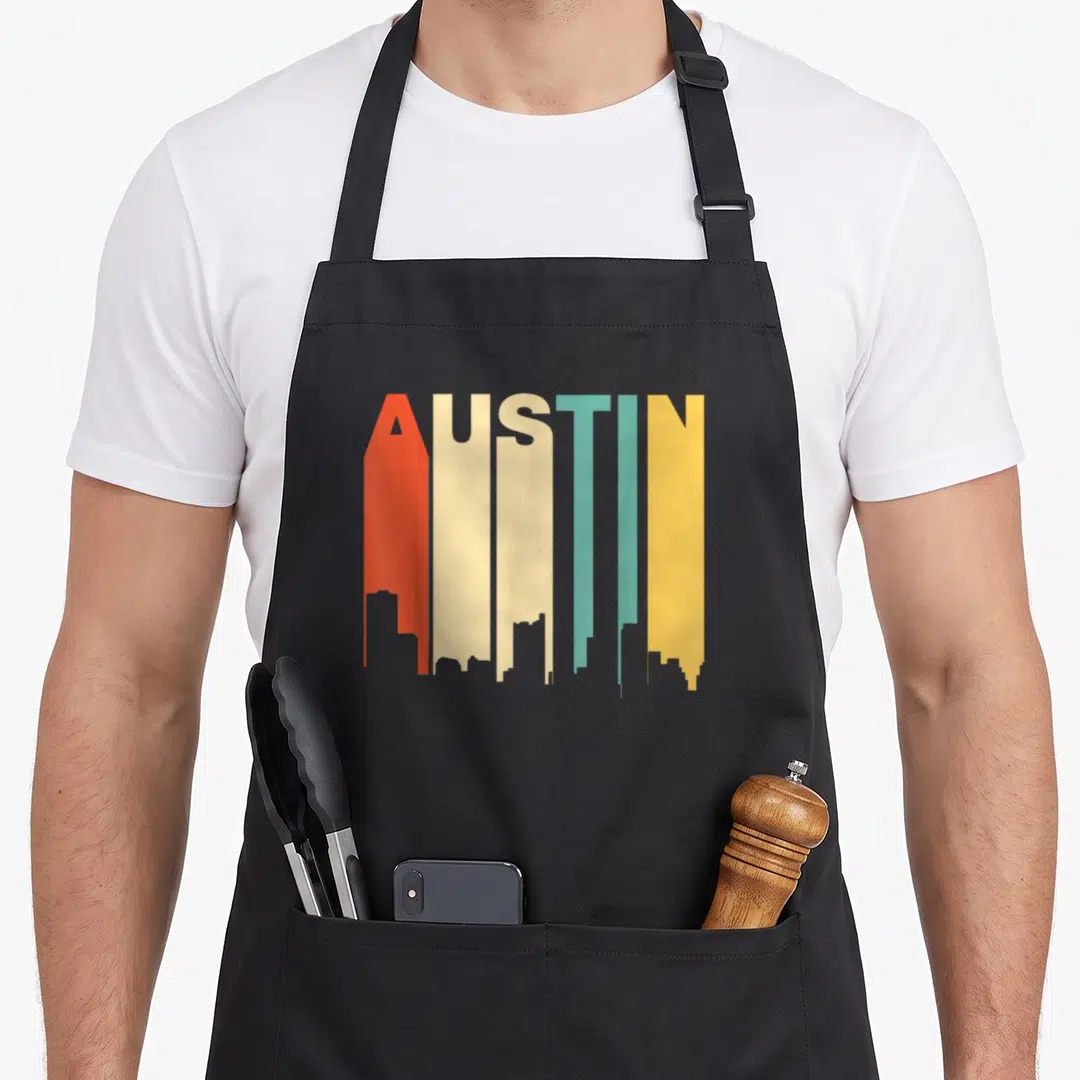 Skyline texas aprons - AUSTIN text forming a colorful city skyline silhouette in orange cream teal and yellow — shown on a black cotton canvas apron against a white background