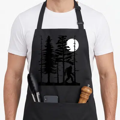 Hiding bigfoot apron - Black silhouette of bigfoot walking among pine trees under a white moon in line art style — shown on a black cotton canvas apron against a white background