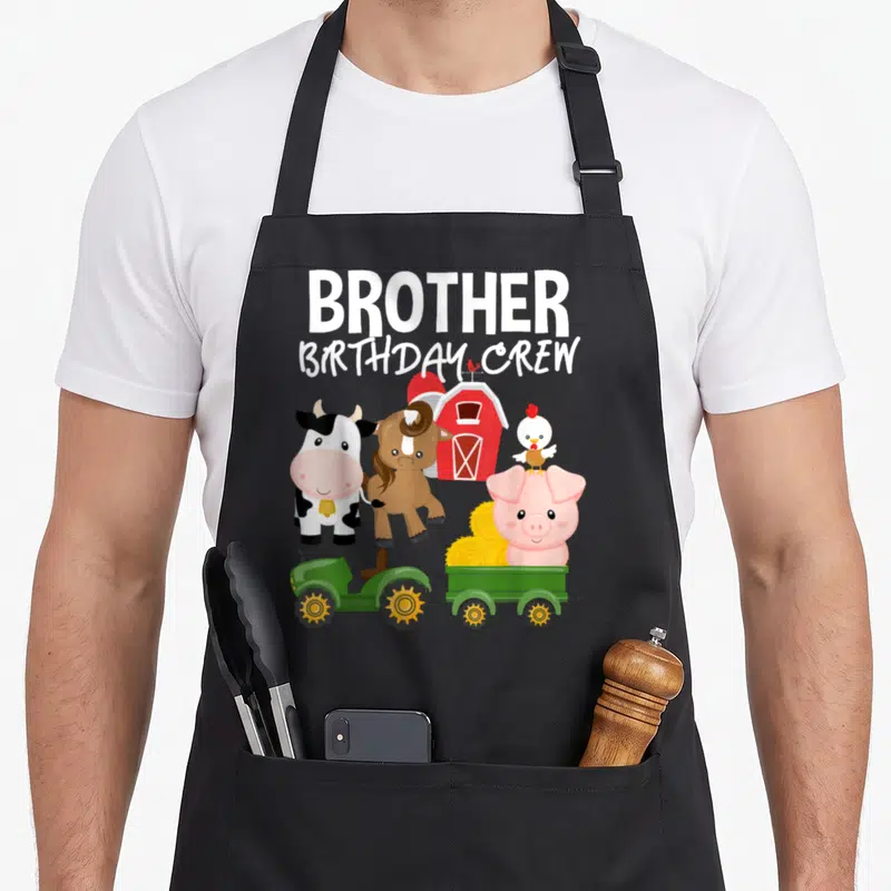 Brother farm aprons - Stacked Brother Birthday Crew text above cartoon cow, horse, pig, chicken, red barn, and green — shown on a black cotton canvas apron against a white background