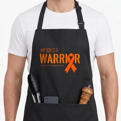 Son leukemia awareness apron - Orange text My Son Is A Warrior with orange ribbon overlapping the letter O — shown on a black cotton canvas apron against a white background