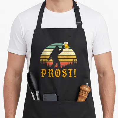 Prost yeti apron - Bigfoot silhouette holding beer mug against retro striped sun with PROST text — shown on a black cotton canvas apron against a white background