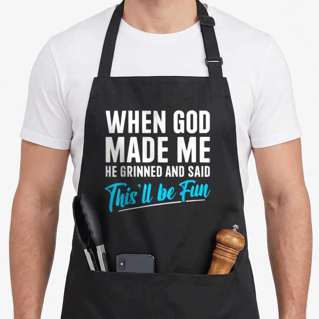 God funny christian apron - Stacked white and blue text reading When God Made Me He Grinned and Said This'll be Fun — shown on a black cotton canvas apron against a white background