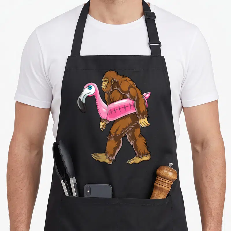 Pool pink flamingo apron - Brown bigfoot walking left carrying pink flamingo floatie cartoon art — shown on a black cotton canvas apron against a white background