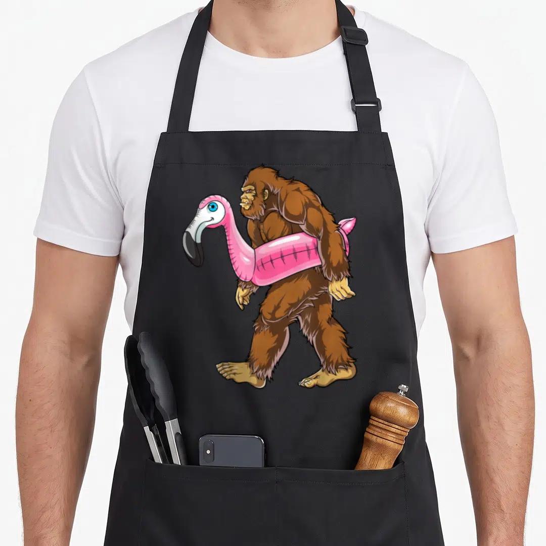 Pool pink flamingo apron - Brown bigfoot walking left carrying pink flamingo floatie cartoon art — shown on a black cotton canvas apron against a white background