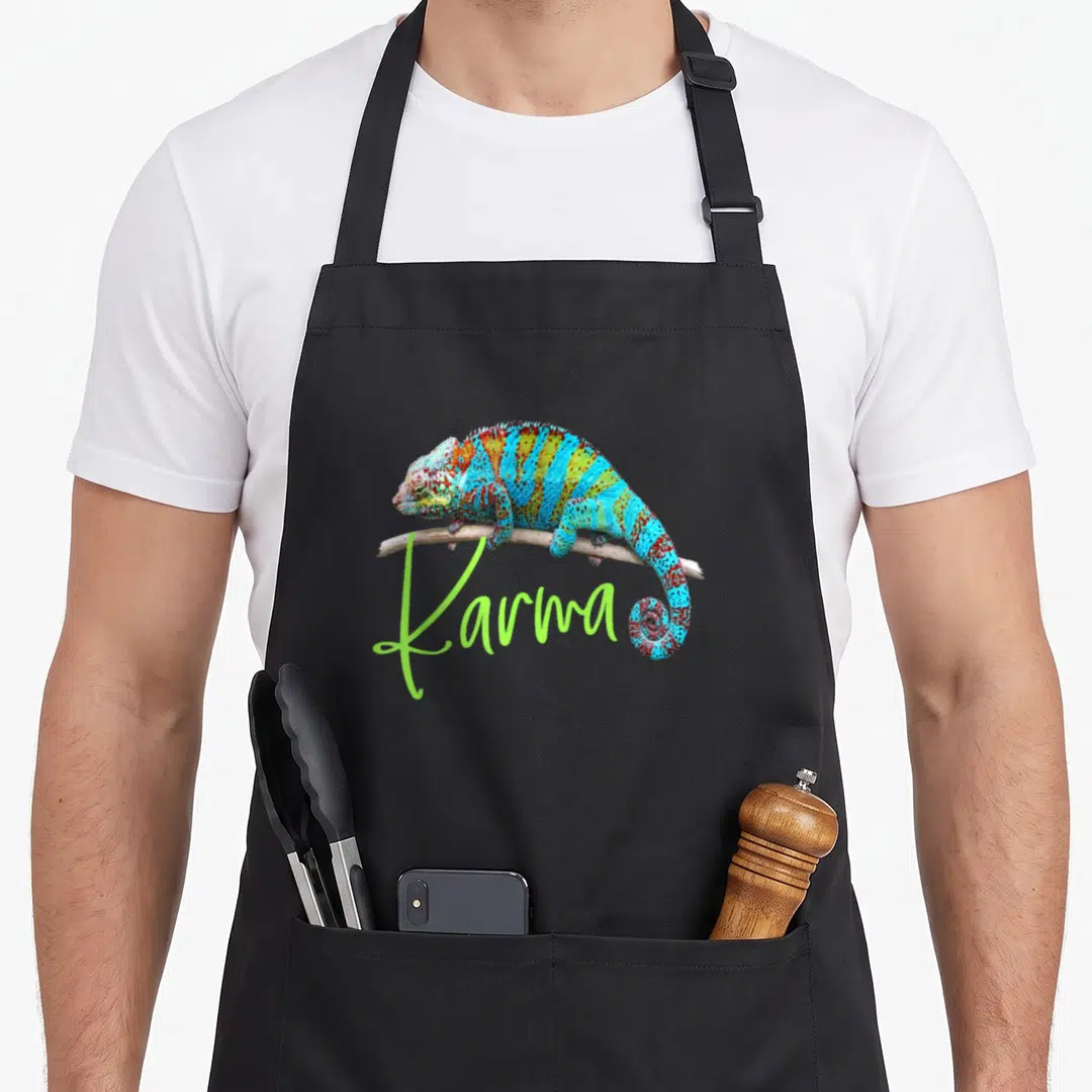 Chameleon 1980s apron - Colorful chameleon perched on a branch with large lime green Karma script text overlapping — shown on a black cotton canvas apron against a white background
