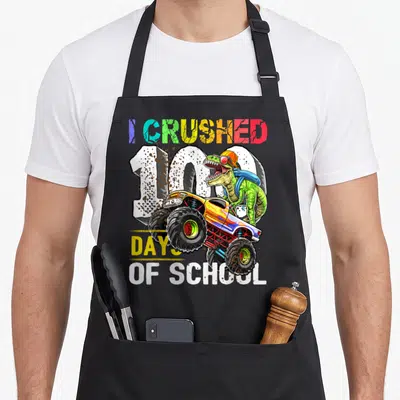 Crushed school truck apron - Green dinosaur driving monster truck with I Crushed 100 Days of School text — shown on a black cotton canvas apron against a white background