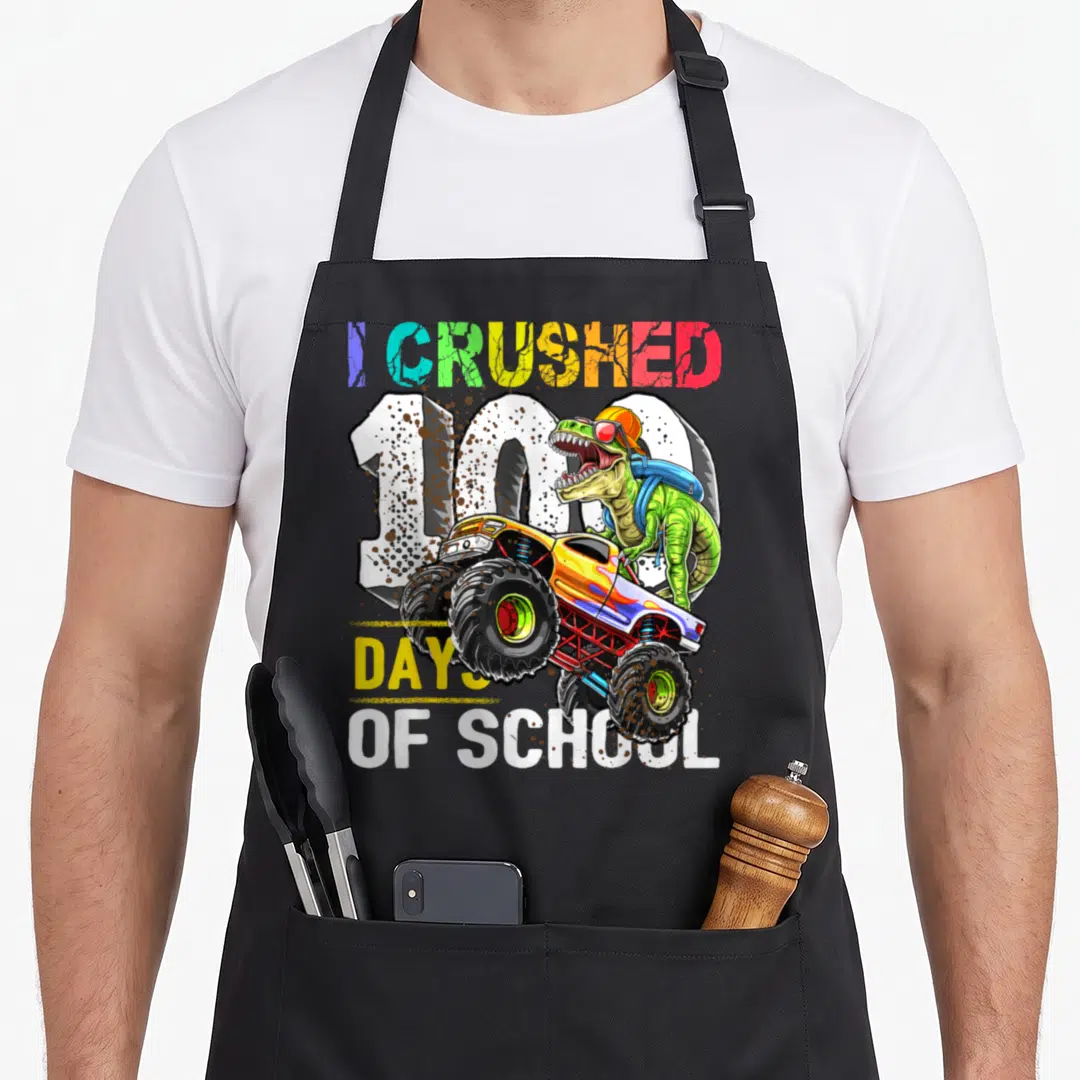 Crushed school truck apron - Green dinosaur driving monster truck with I Crushed 100 Days of School text — shown on a black cotton canvas apron against a white background