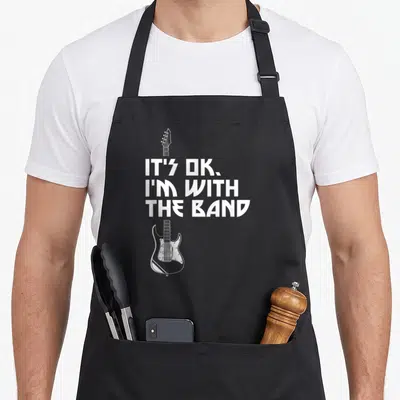 Band roadie apron - White stacked text IT'S OK I'M WITH THE BAND flanking a white bass guitar outline on black fabric — shown on a black cotton canvas apron against a white background