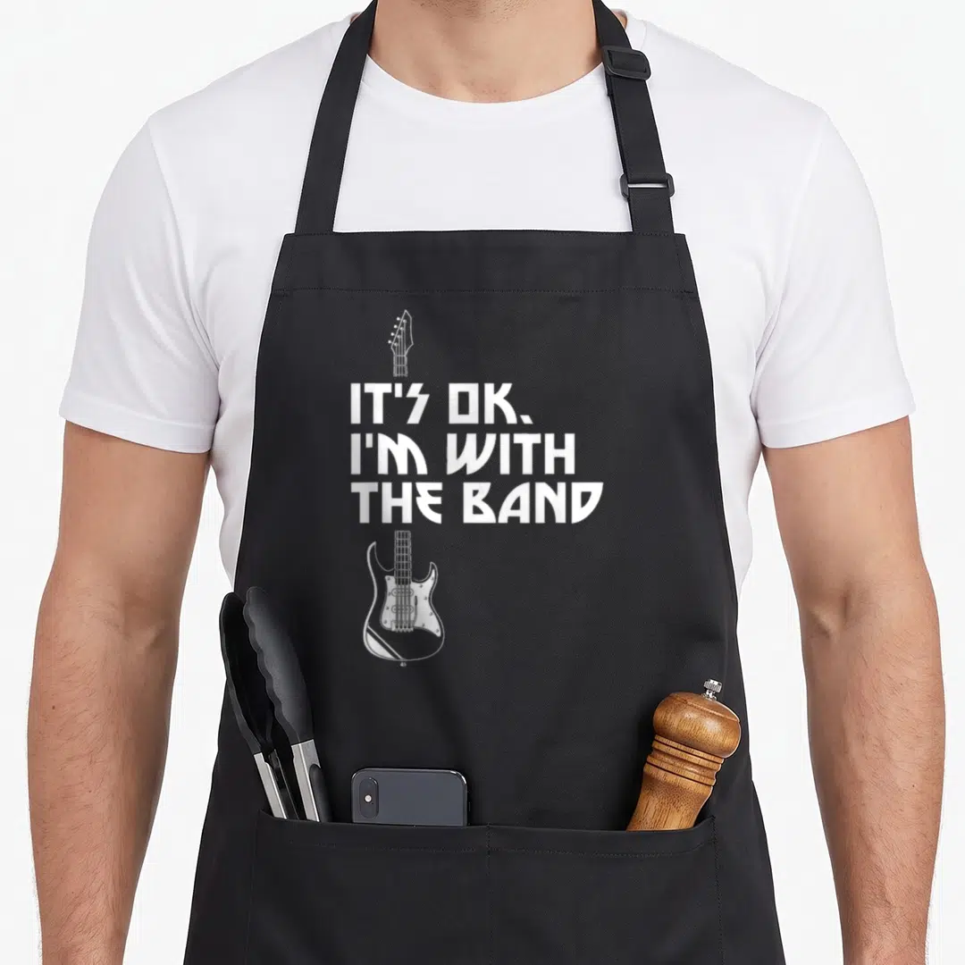 Band roadie apron - White stacked text IT'S OK I'M WITH THE BAND flanking a white bass guitar outline on black fabric — shown on a black cotton canvas apron against a white background