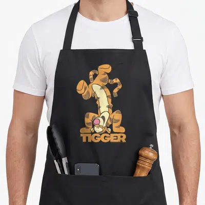 Tigger novelty apron - Upside down cartoon tiger in orange and tan with bold TIGGER text below — shown on a black cotton canvas apron against a white background