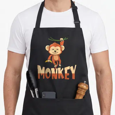 Zoo toddler animal apron - Cartoon brown monkey hanging from green vine holding yellow banana above two-tone MONKEY text — shown on a black cotton canvas apron against a white background