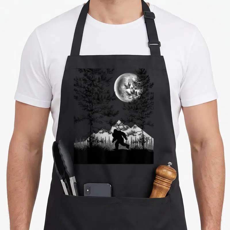 Bigfoot walking apron - Sasquatch silhouette between two pine trees under a full moon in black and grey — shown on a black cotton canvas apron against a white background
