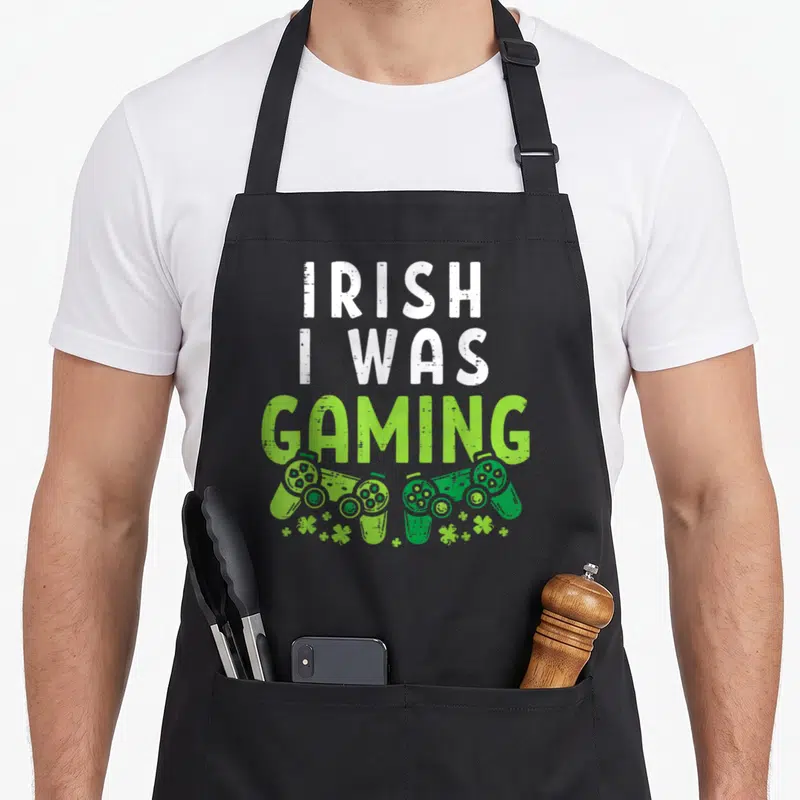 Gaming irish aprons - Stacked IRISH I WAS GAMING text above two green controllers and small shamrocks — shown on a black cotton canvas apron against a white background