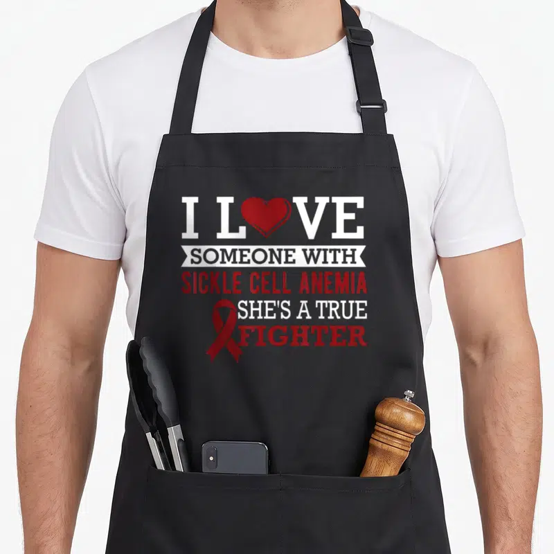 Cell awareness ribbon apron - Red and white text I Love Someone With Sickle Cell Anemia She's A True Fighter with ribbon — shown on a black cotton canvas apron against a white background