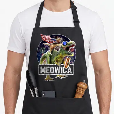 Meowica 4th of july apron - Green T-Rex holding hot dog with cat in flag cape and MEOWICA text over fireworks — shown on a black cotton canvas apron against a white background