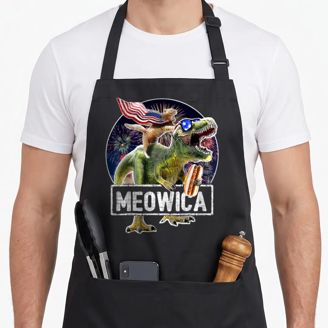 Meowica 4th of july apron - Green T-Rex holding hot dog with cat in flag cape and MEOWICA text over fireworks — shown on a black cotton canvas apron against a white background