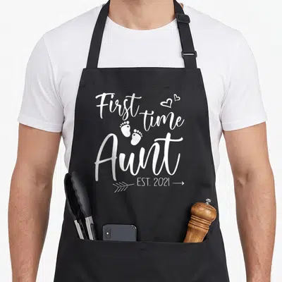 Aunt christmas aprons - White text First time Aunt EST 2021 with two hearts and baby footprints — shown on a black cotton canvas apron against a white background