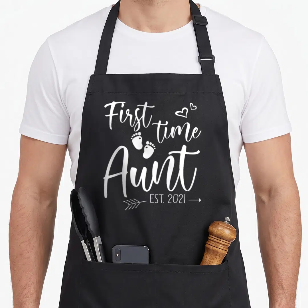 Aunt christmas aprons - White text First time Aunt EST 2021 with two hearts and baby footprints — shown on a black cotton canvas apron against a white background