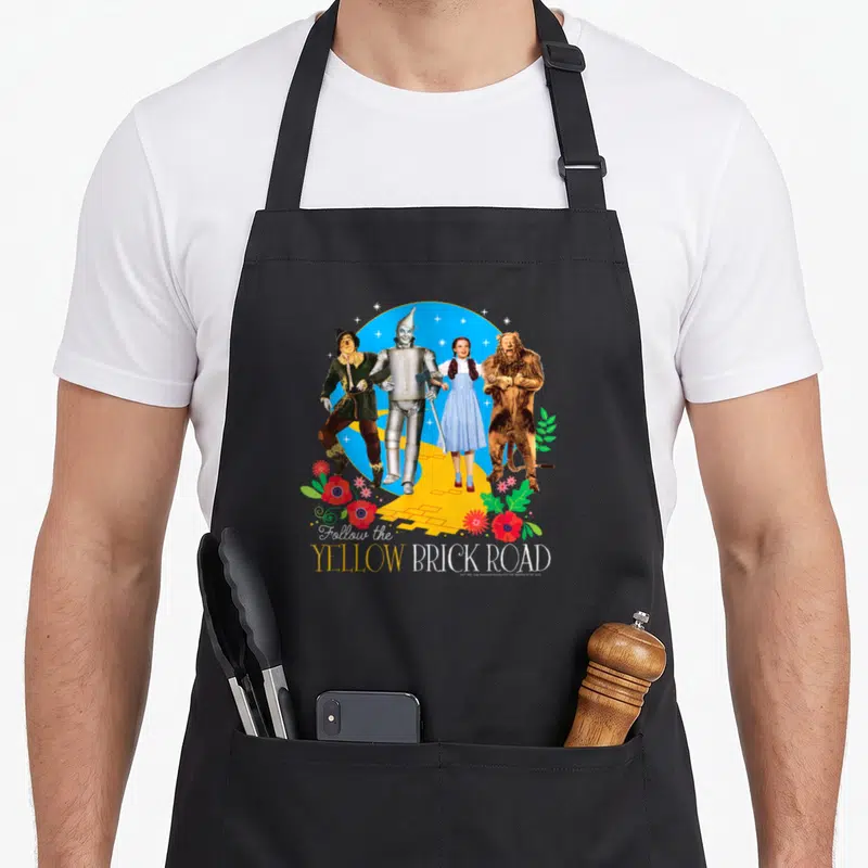 Wizard road apron - Four characters on yellow brick road with red flowers and Follow the YELLOW BRICK ROAD text — shown on a black cotton canvas apron against a white background