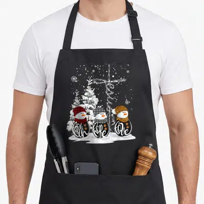Hope christmas snowman apron - Three snowmen with Faith Hope Love text, plaid and leopard hats, snowflake cross — shown on a black cotton canvas apron against a white background
