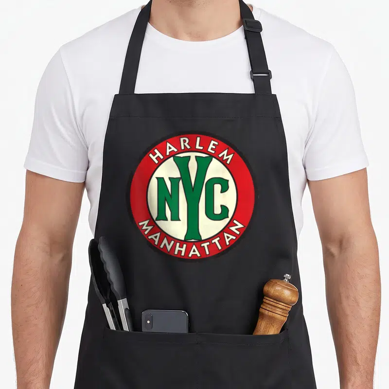 Manhattan nyc apron - Circular red emblem with HARLEM NYC MANHATTAN text in green and white — shown on a black cotton canvas apron against a white background