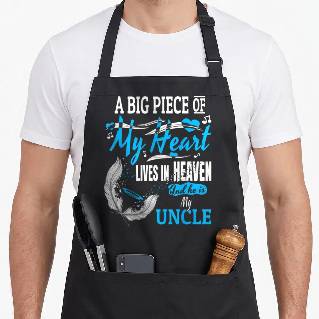 Live uncle life apron - Stacked text A Big Piece Of My Heart Lives In Heaven with white feather and blue musical notes — shown on a black cotton canvas apron against a white background