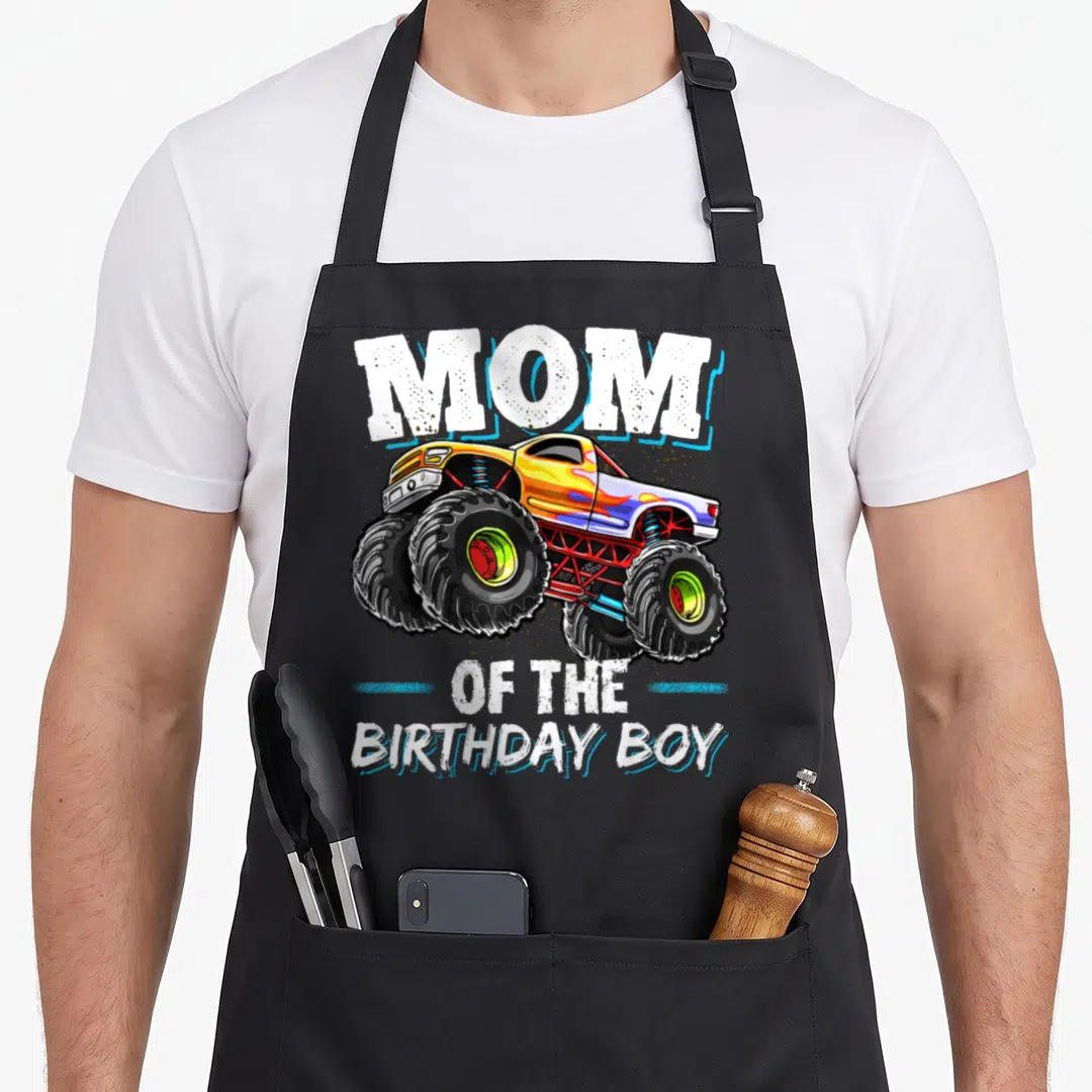 Birthday monster truck apron - MOM OF THE BIRTHDAY BOY text with yellow and blue monster truck illustration — shown on a black cotton canvas apron against a white background